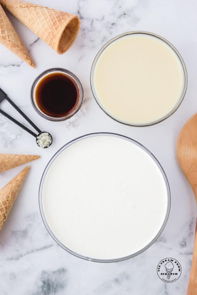 Ingredients needed to make no churn vanilla ice cream in separate bowls on a marble counter along with sugar cones.