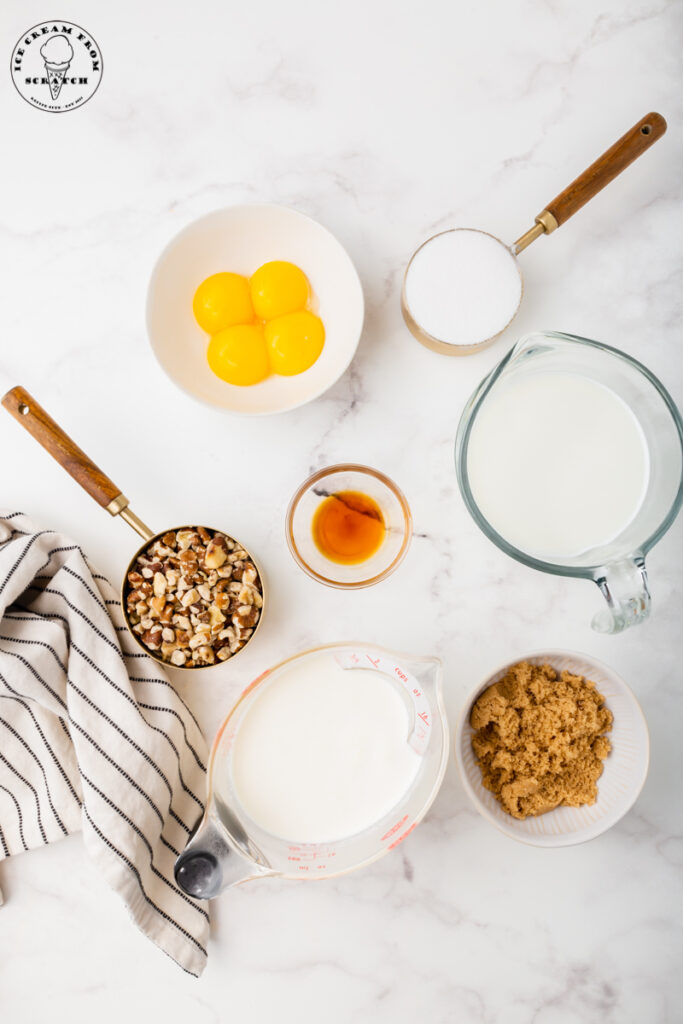 ingredients needed for making black walnut ice cream, including nuts, sugar, milk, eggs, and brown sugar, each in separate dishes on a marble countertop
