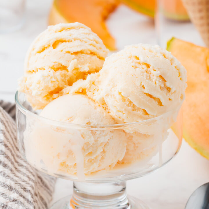 four scoops of melon ice cream in a glass ice cream dish. There are slices of cantaloupe in the background.