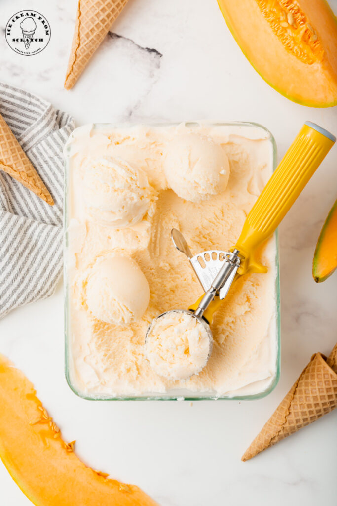 a rectagular glass dish filled with melon ice cream. Four scoops have been made with a yellow handled ice cream scoop. Around the dish are wedges of cantaloupe and sugar cones.