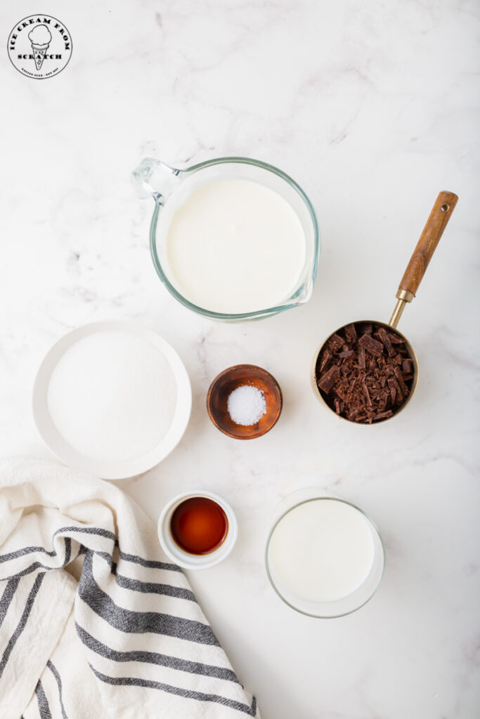 ingredients for homemade chocolate chip ice cream are in separate bowls on a marble counter. Includes chopped chocolate, sugar, and cream.