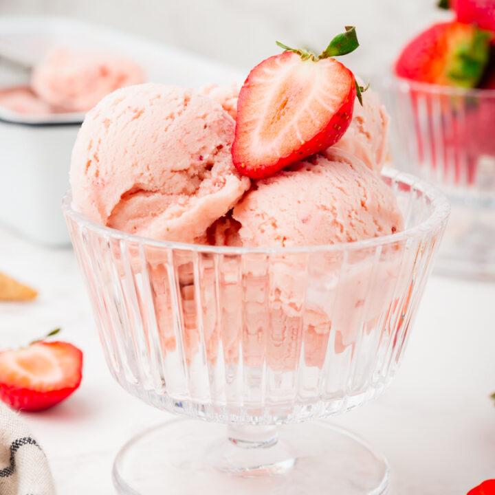 a footed glass dessert dish is filled with homemade strawberry ice cream and topped with half of a fresh strawberry.