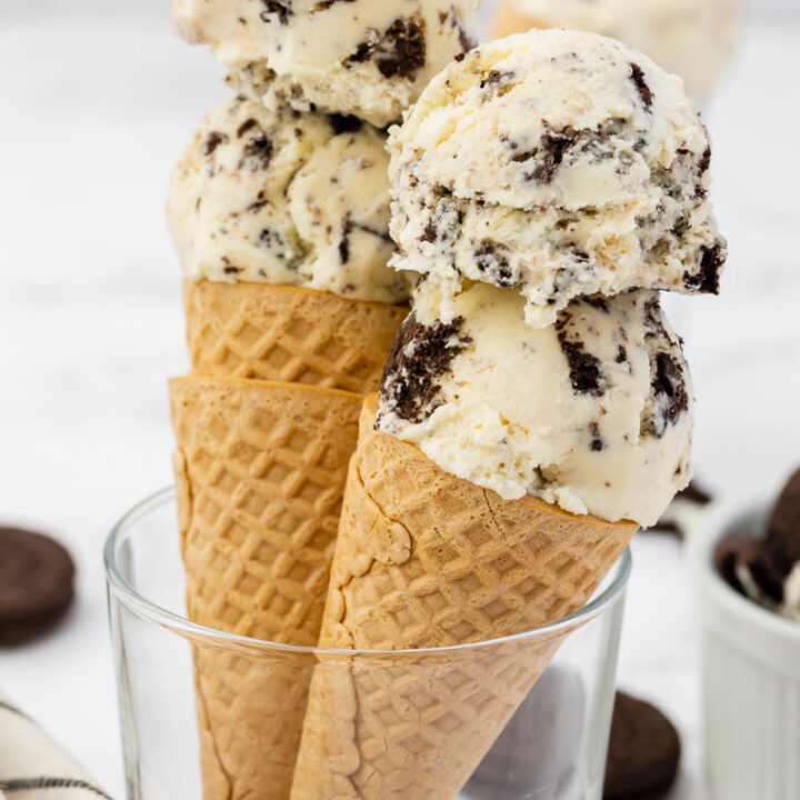 Two sugar cones with double scoops of Oreo cookies and cream ice cream held up in a glass