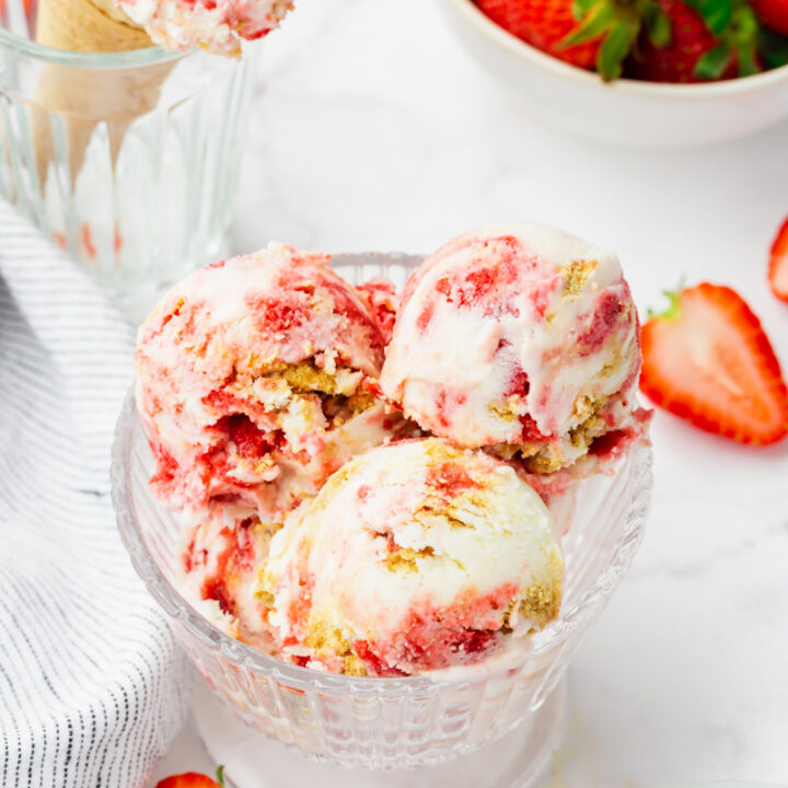 a glass dish filled with three scoops of strawberry cheesecake ice cream packed with berries and graham cracker pieces. In the background is a bowl of strawberries and a cone of the same ice cream.