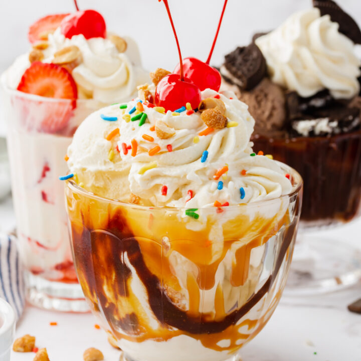 A glass bowl filled with a chocolate caramel ice cream sundae, topped with whipped cream, sprinkles, and two maraschino cherries. More ice cream sundaes of different varieties are in the background.