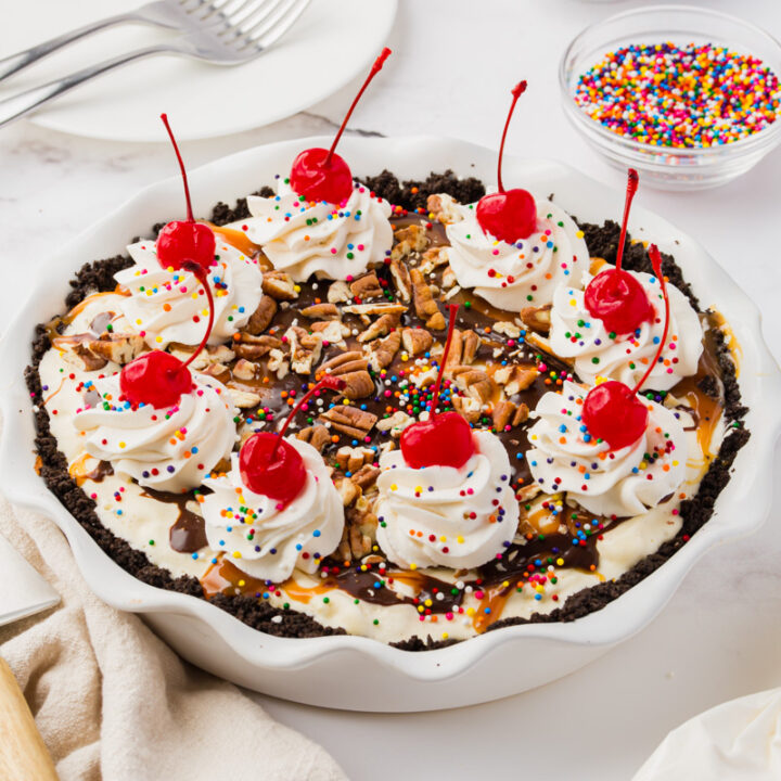 a homemade ice cream pie topped with piles of whipped cream, cherries, sprinkles, and pecans.