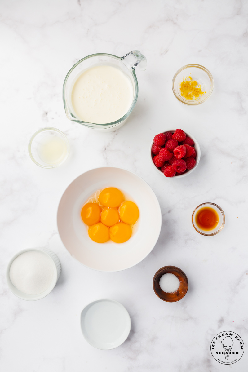 The ingredients needed to make semifreddo from scratch, with raspberries. All of the ingredients are measured into small bowls and arranged on a marble countertop.
