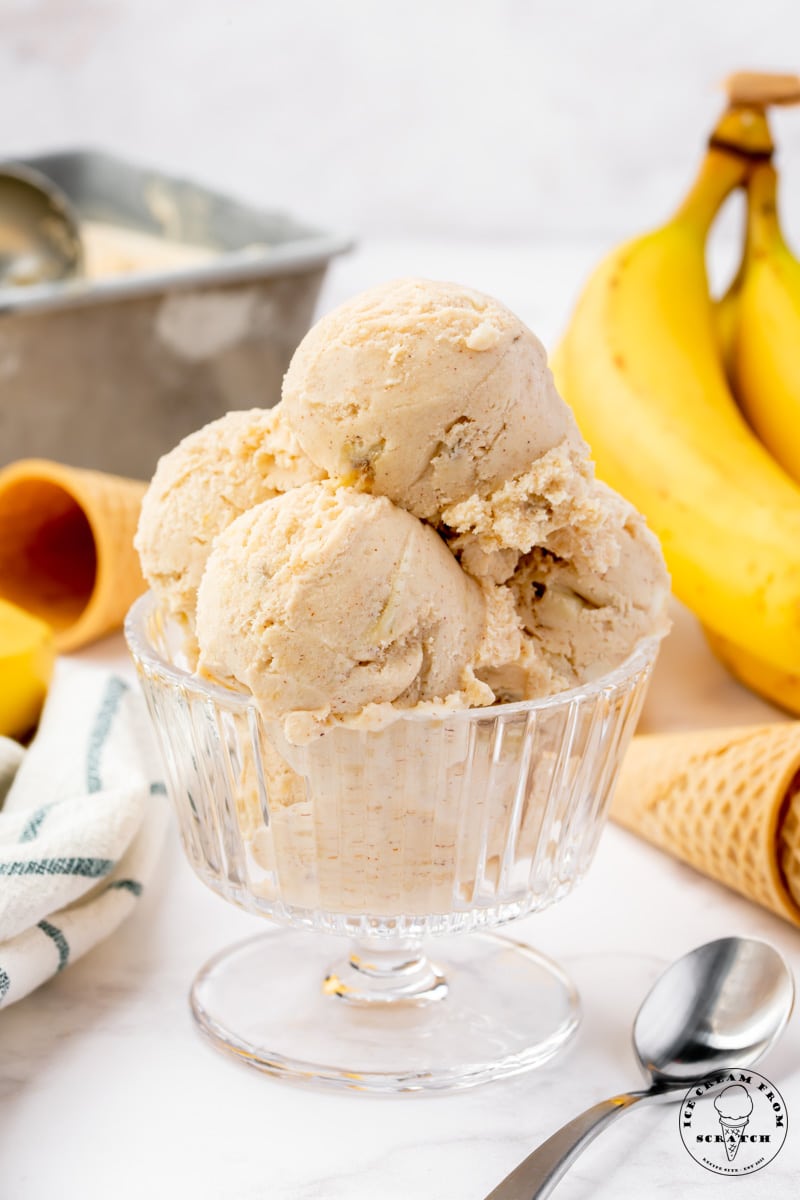 scoops of homemade banana foster ice cream in a footed glass dish, garnished with a slice of banana. Ice Cream cones and yellow bananas are in the background.