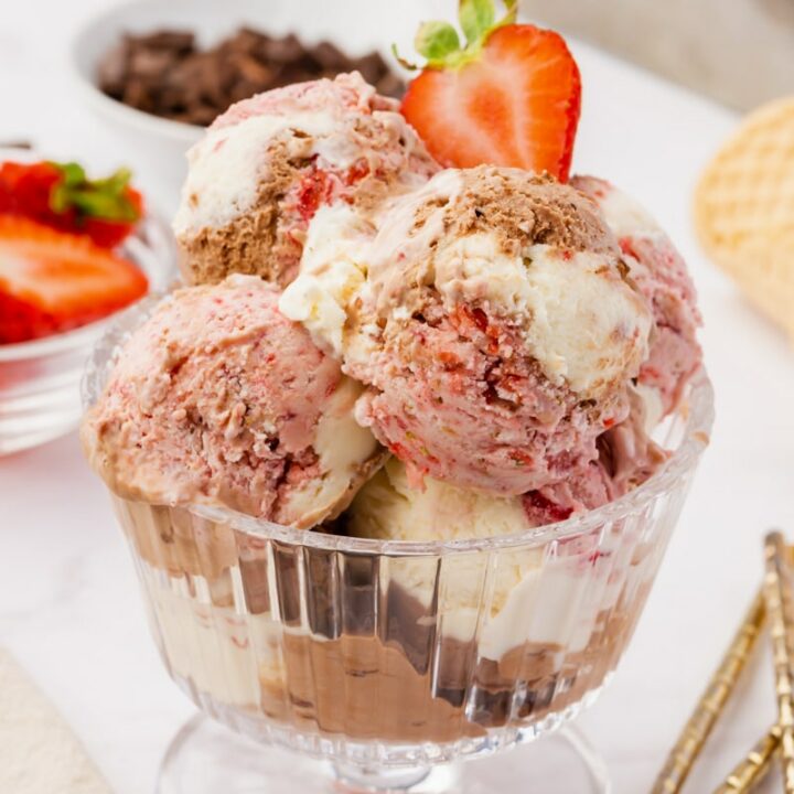 homemade no churn neapolitan ice cream, scooped into a glass, ridged, footed ice cream dish. Two spoons are on the table next to the glass. Dishes of sliced strawberries and chocolate chips are in the background