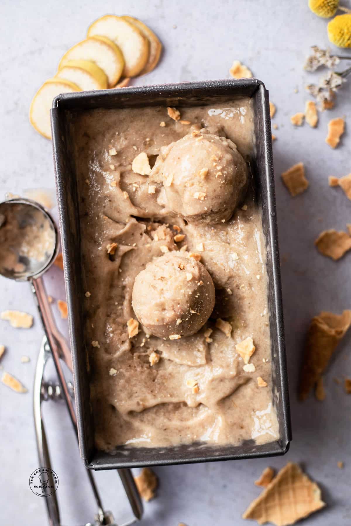 a metal loaf pan filled with banana nice cream. Two scoops have been made and placed on top. Crumbled sugar cones are sprinkled over and around the pan.