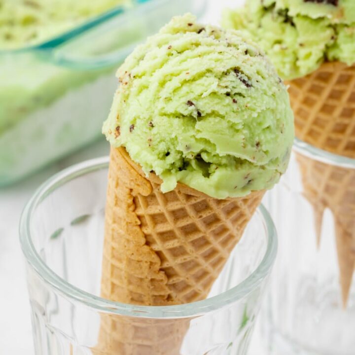 coconut mint ice cream scooped into two sugar cones that are propped up in glasses so that they are upright. The ice cream is green with chocolate chunks in it.