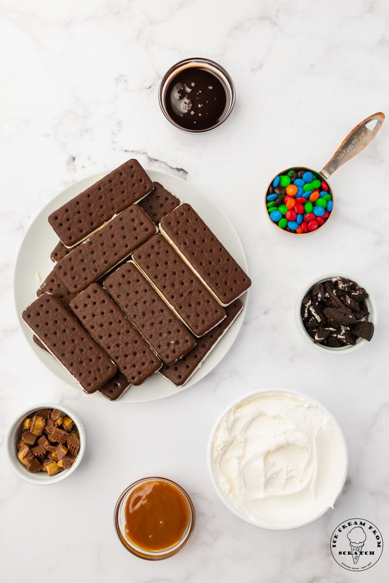 A plate of ice cream sandwiches. Around the plate are small bowls filled with chopped oreos, chopped peanut butter cups, chocolate sauce, and caramel sauce. A larger bowl holds whipped topping, and a measuring cup holds m&m candy.