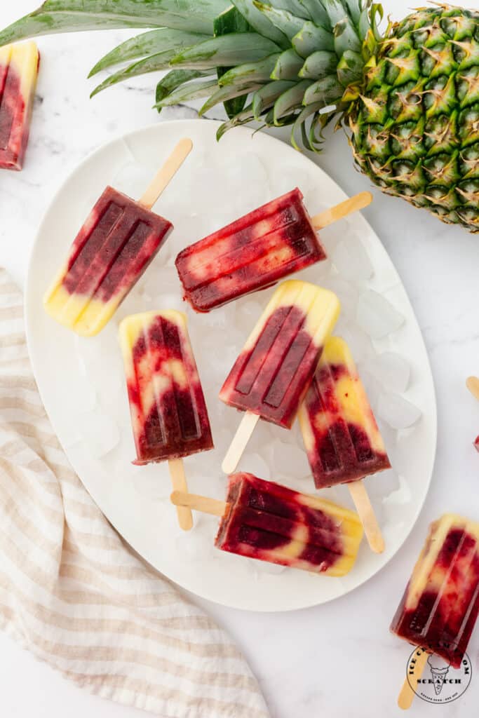 8 swirled cherry pineapple popsicles on a tray of ice, viewed from above. The tray is next to a whole fresh pineapple.