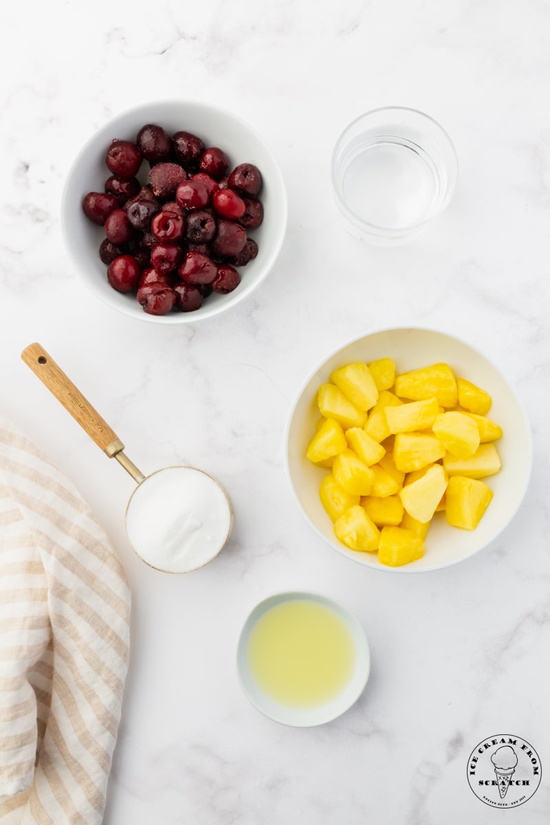 The ingredients for cherry pineapple popsicles (frozen pineapple, frozen cherries, sugar, lemon juice, and water) each in separate bowls on the counter, viewed from overhead.