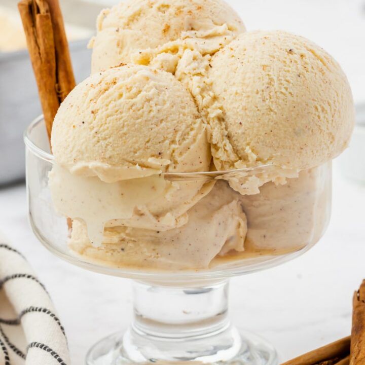 A footed glass dish holding 5 scoops of homemade eggnog ice cream, speckled with cinnamon and spices. There are rolled cinnamon sticks on the counter next to the dish, and stuck into the ice cream.