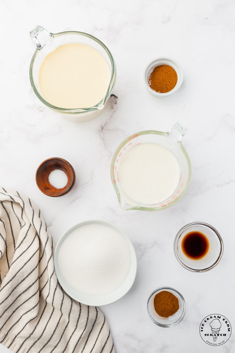 A marble counter top with a glass measuring cup of cream, a glass measuring cup of milk, a bowl of sugar, and small bowls with the flavorings for eggnog ice cream.