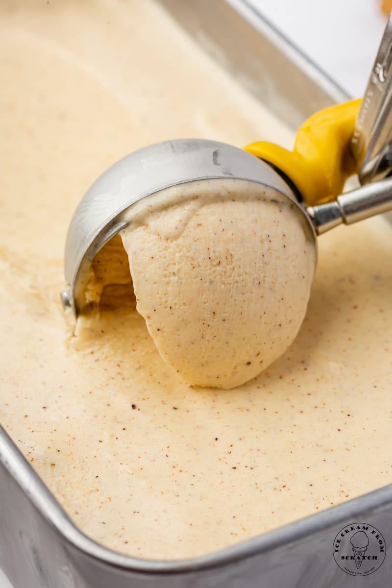 a metal loaf pan of homemade eggnog ice cream. A metal ice cream scoop is creating a serving.