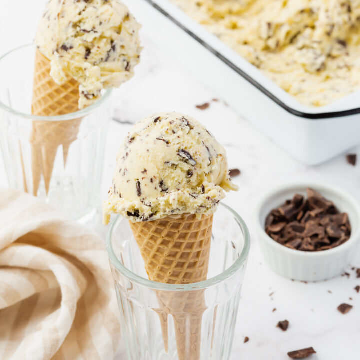 Two cones of homemade stracciatella ice cream held up in glasses, next to a white loaf pan filled with ice cream.
