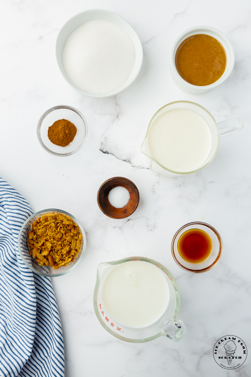 The ingredients needed to make cookie butter ice cream from scratch, all in small bowls, arranged on a marble counter, and viewed from above.