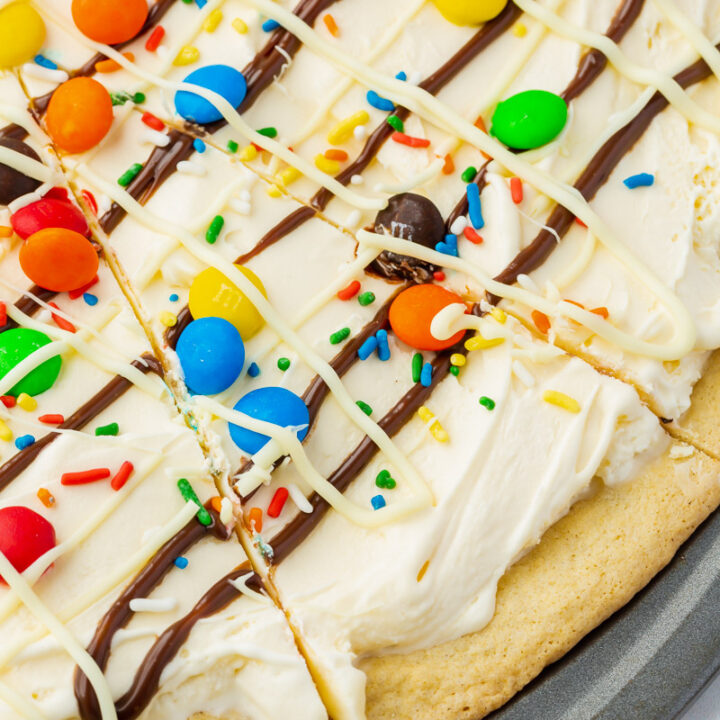 closeup of the edge of a homemade ice cream pizza in a metal pizza pan. the dessert is topped with white chocolate, hot fudge, sprinkles, and m&ms