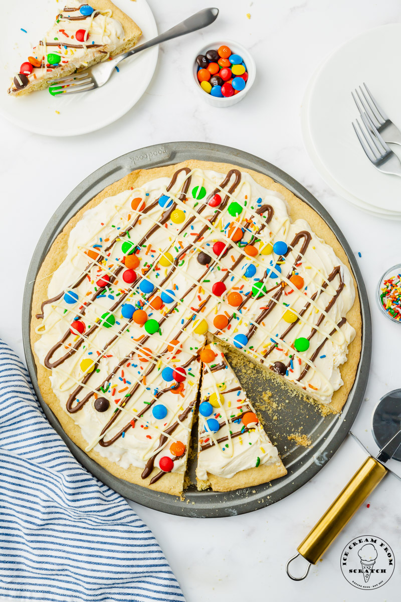 a large round cookie topped with ice cream to create an ice cream pizza with candy, sprinkles, and chocolate toppings.