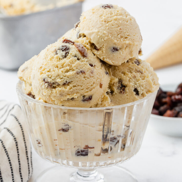 a footed, fluted glass dish filled with scooops of rum raisin ice cream. raisins are on the counter next to the bowl. in the background is an ice cream cone and a metal loaf pan.