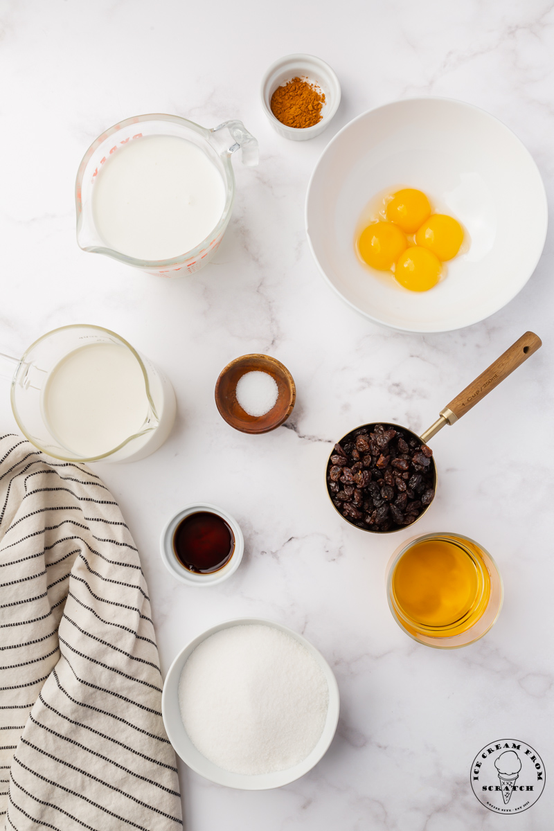 The ingredients needed to make a french-style rum raisin ice cream, all in separate bowls, arranged on a marble counter, and viewed from above.