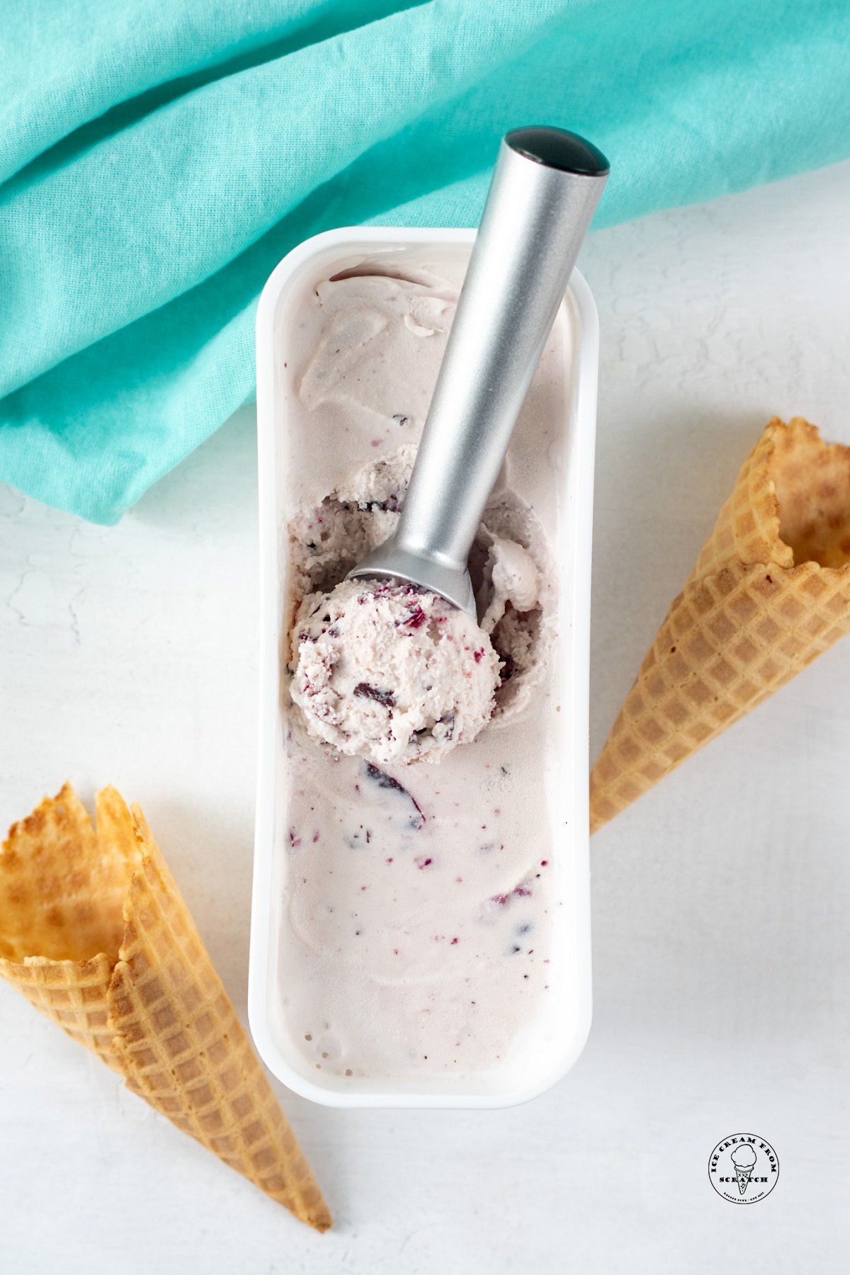 overhead view of a rectangular ice cream container filled with homemade amaretto ice cream. A metal scoop is serving the ice cream, and there are waffle cones on the counter next to the container.