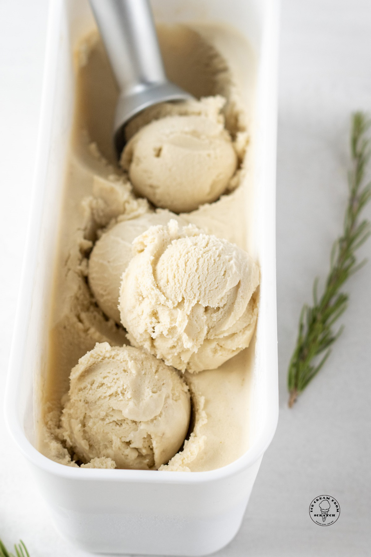scoops of homemade rosemary ice cream in a white plastic rectangular container with a metal ice cream scoop.
