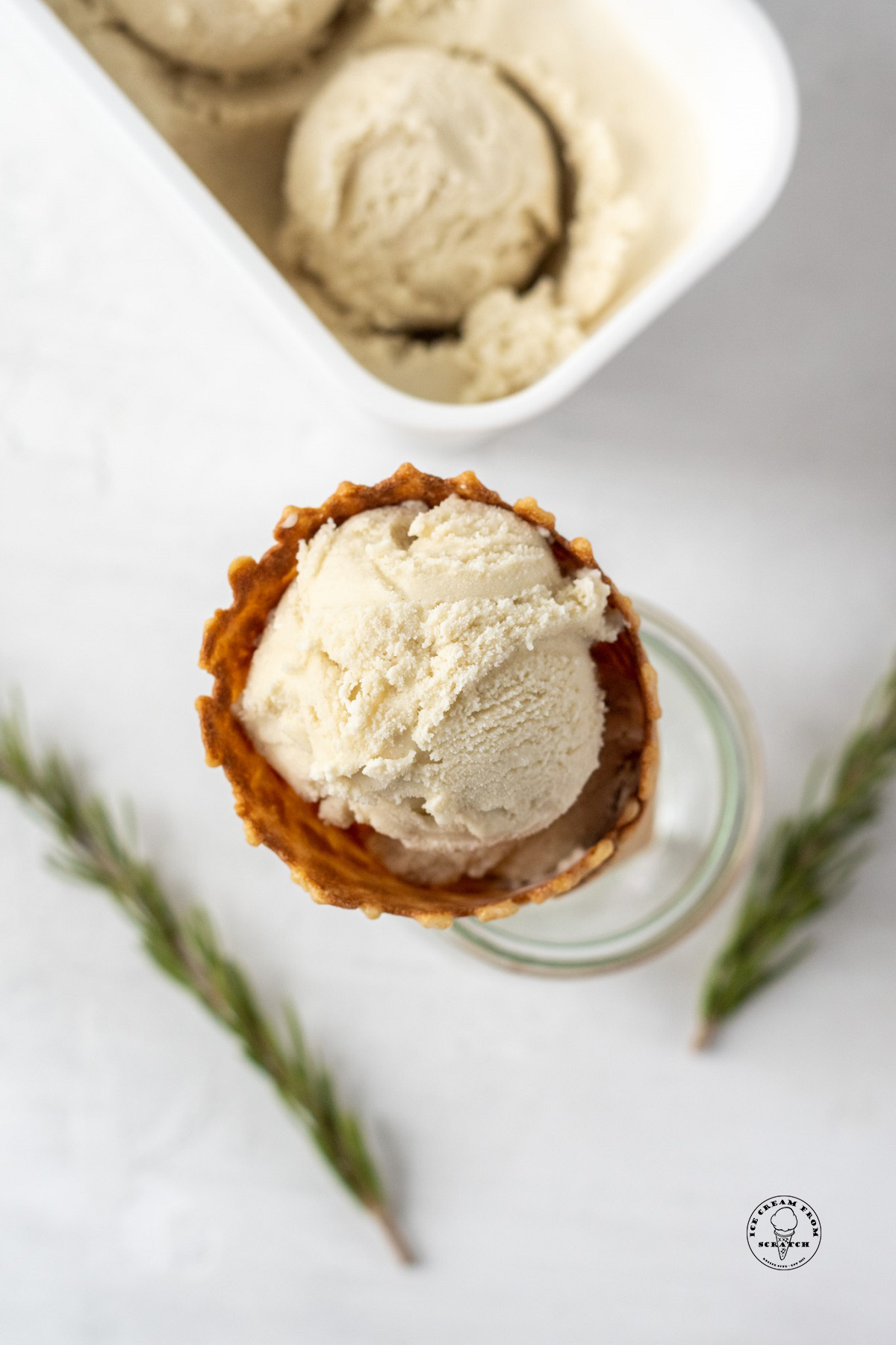 A waffle cone filled with rosemary ice cream, viewed from overhead. In the background are sprigs of fresh herbs.
