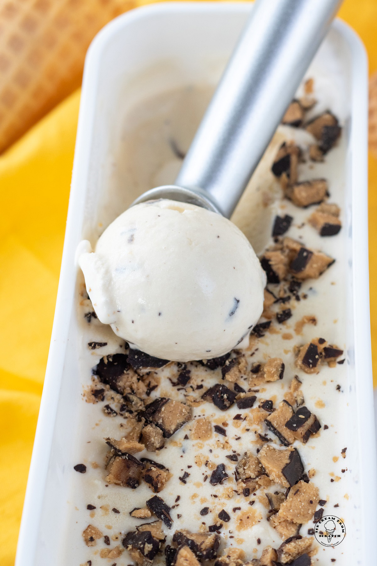 a metal ice cream scoop pulling through a rectangular ice cream container of butter brickle.