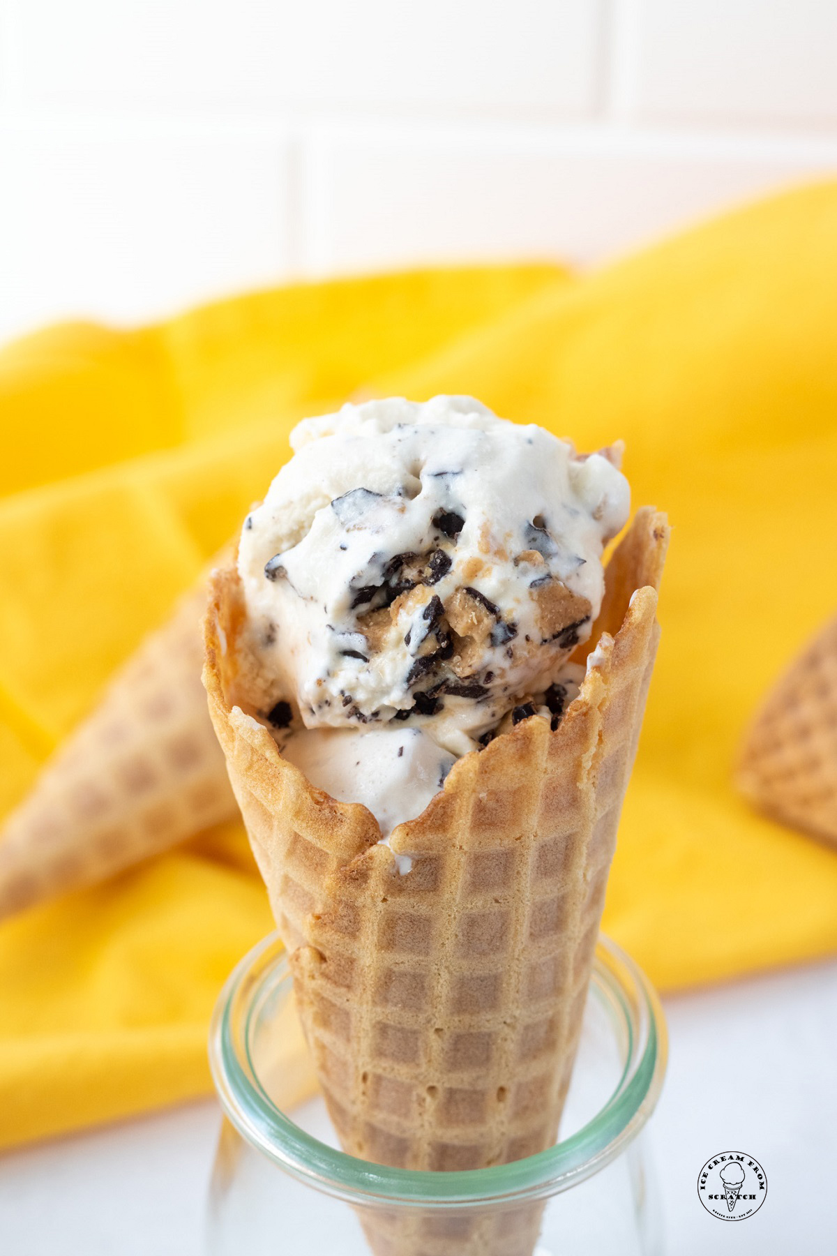 a waffle cone holding homemade ice cream with butter brickle toffee pieces. in the background is a yellow cloth.