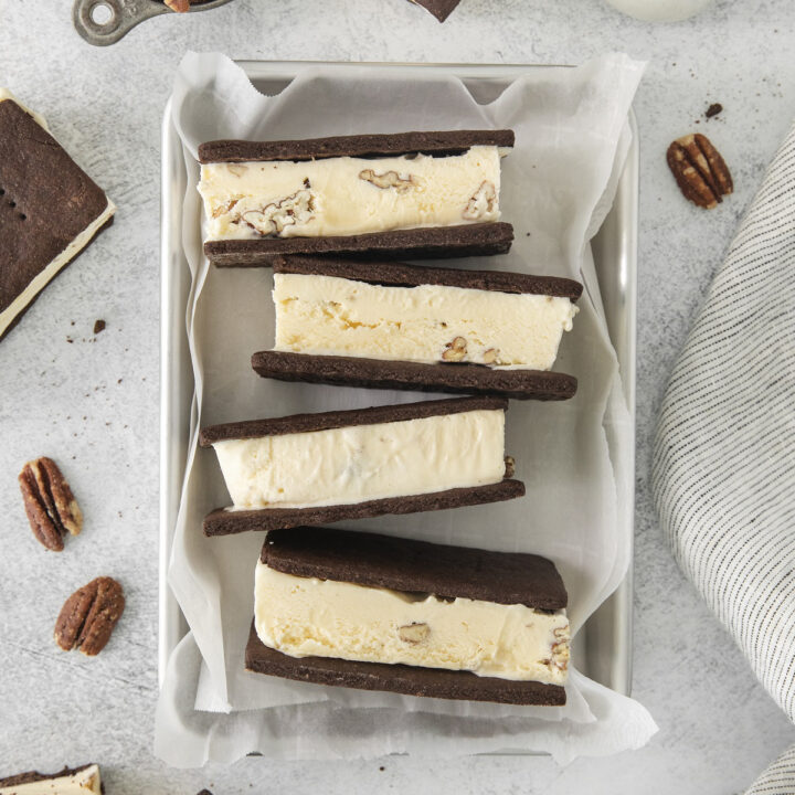 four butter pecan ice cream sandwiches arranged on parchment paper in a baking pan. Around the pan are more sandwiches, a cup of pecans, and a jug of milk.