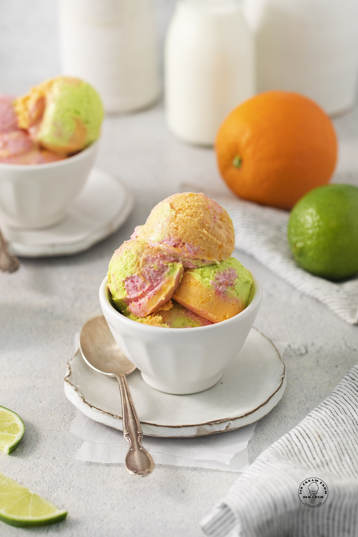 A bowl of scoops of homemade rainbow sherbet in front of bottles of cream, an orange, and a lime.