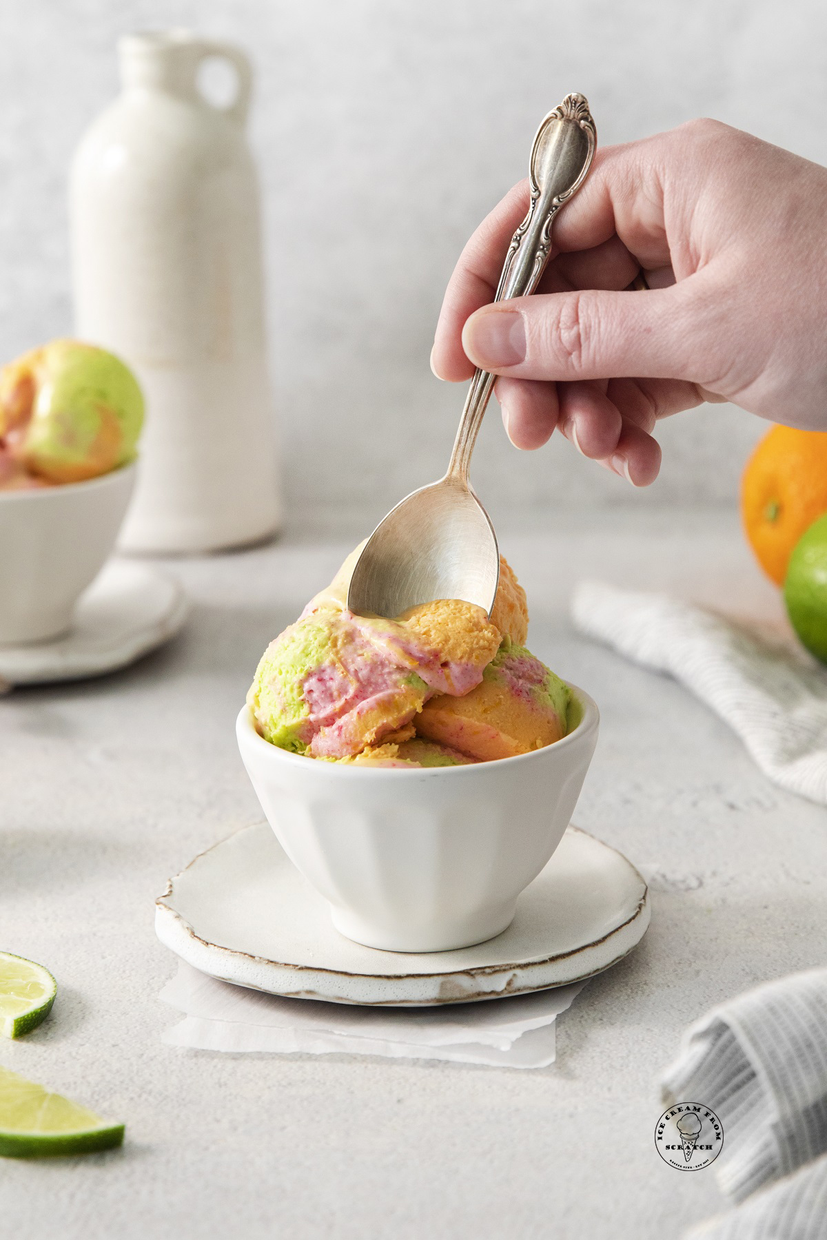 a small white bowl of rainbow sherbet on a ceramic saucer. A hand is eating the sherbet with a silver spoon.