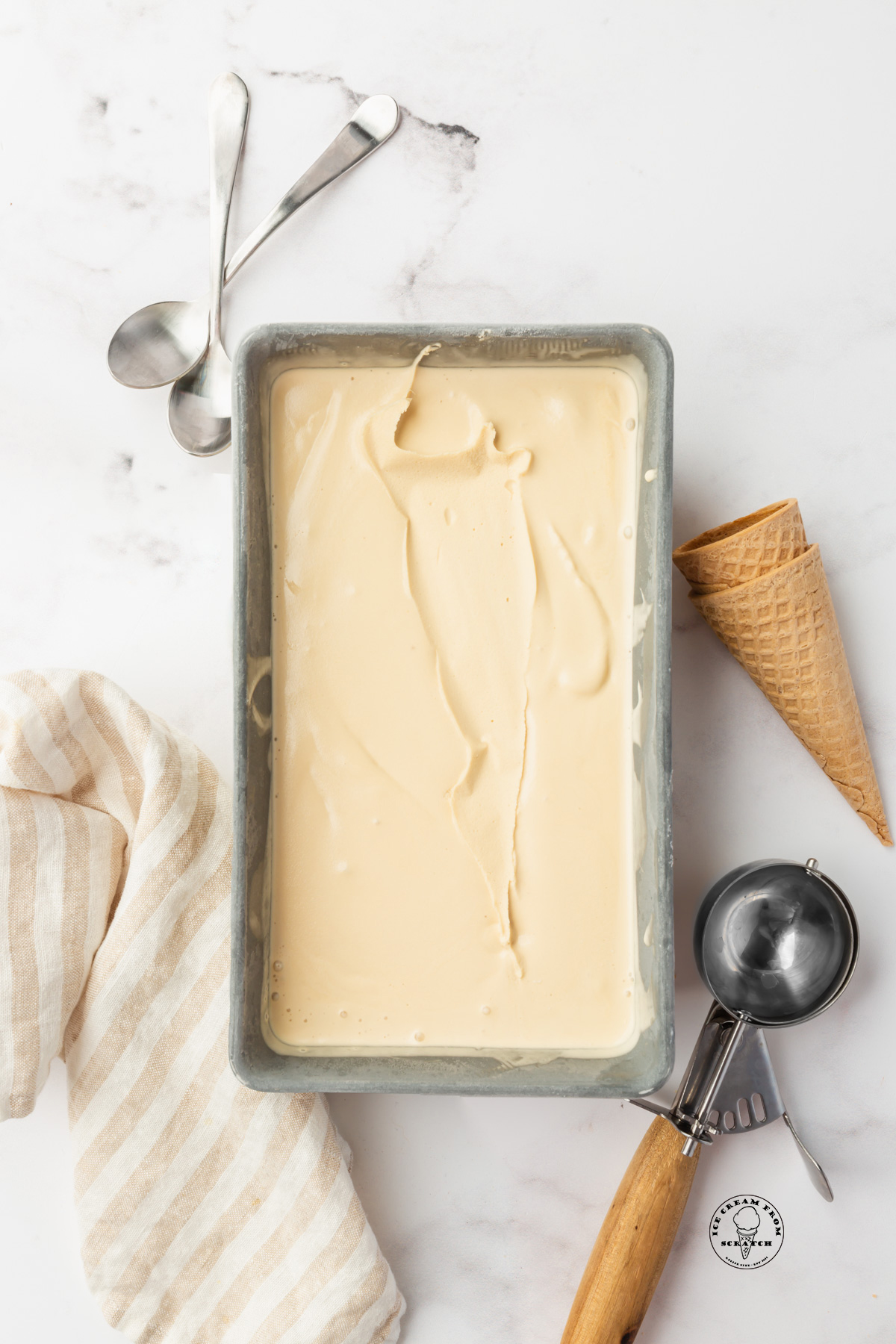 Homemade caramel ice cream in a metal loaf pan. an ice cream scoop and sugar cones are next to the pan.