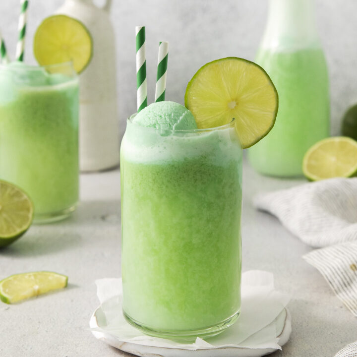 a can shaped glass filled with lime sherbet punch. It's garnished with lime wedges and two green and white straws.