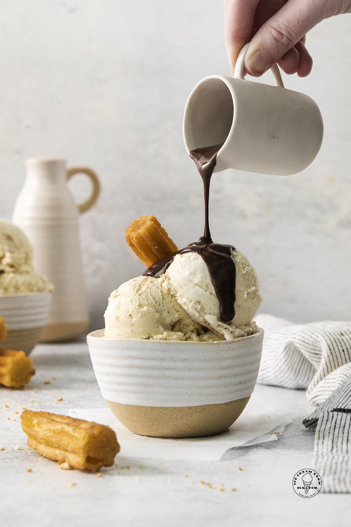 a bowl of churro ice cream with a churro stuck into it and chocolate sauce being poured over.