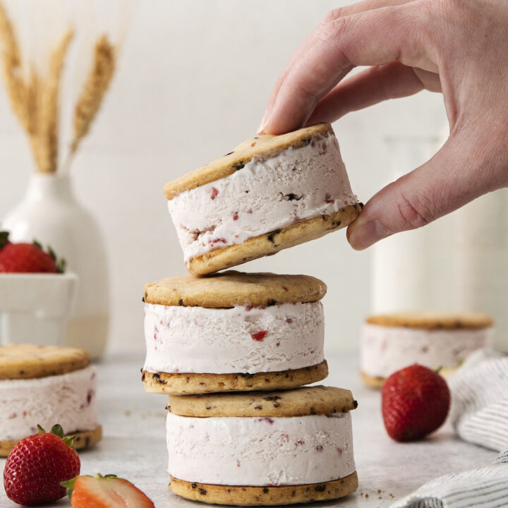 three round strawberry cookie ice cream sandwiches in a stack. A hand is picking one up.