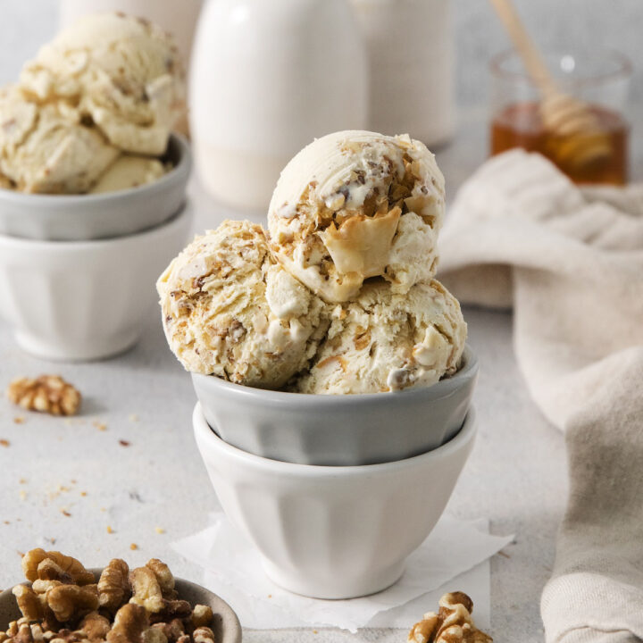 a white and grey ceramic bowl holding scoops of homemade baklava ice cream. There is a small bowl of walnuts in the counter and a cup of honey in the background.