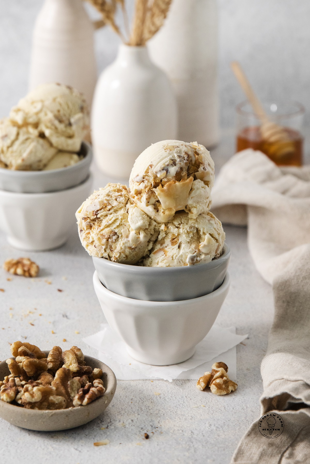 a white and grey ceramic bowl holding scoops of homemade baklava ice cream. There is a small bowl of walnuts in the counter and a cup of honey in the background.