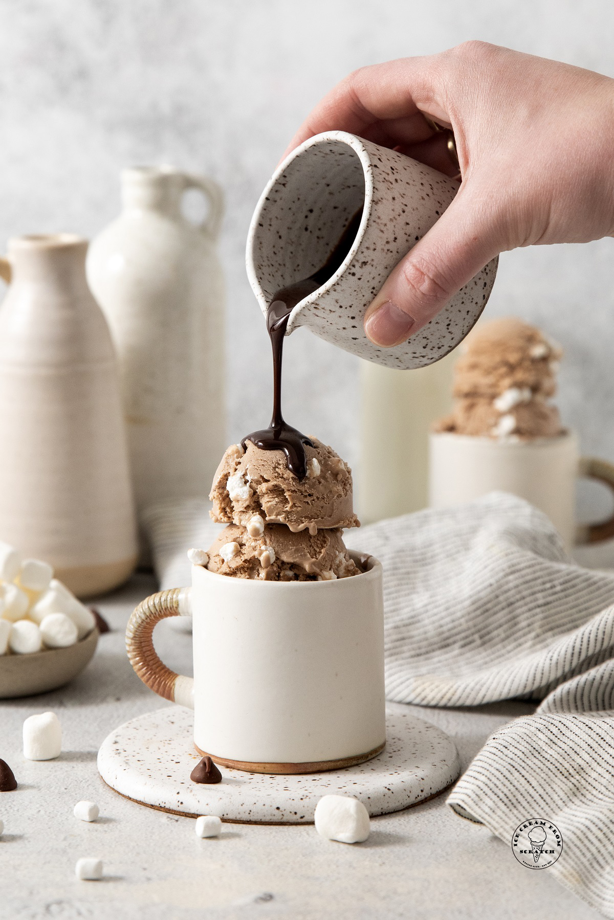 a hand pouring hot fudge from a small white and brown speckled pitcher over a mug filled with hot chocolate ice cream.
