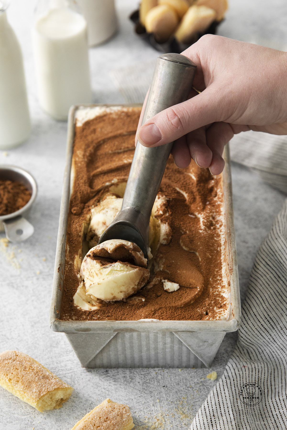 a metal bread pan filled with tiramisu ice cream, dusted with cocoa powder. A metal scoop is pulling up a serving.